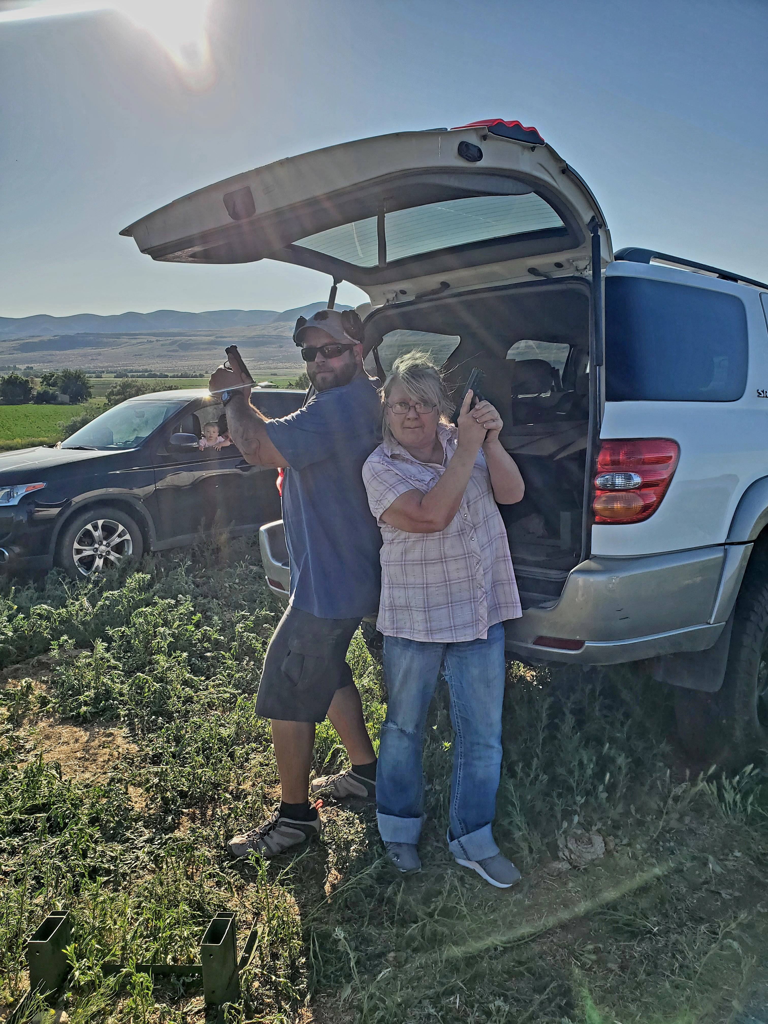 Beef tallow maker's husband and mom holding firearms standing next to an open car trunk in a field.