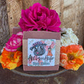 A maroon colored bar of soap, with a rose-gold sparkly wrap, with Rosy Cheeks being the name of the soap. The beef tallow soap also has fresh roses around it.