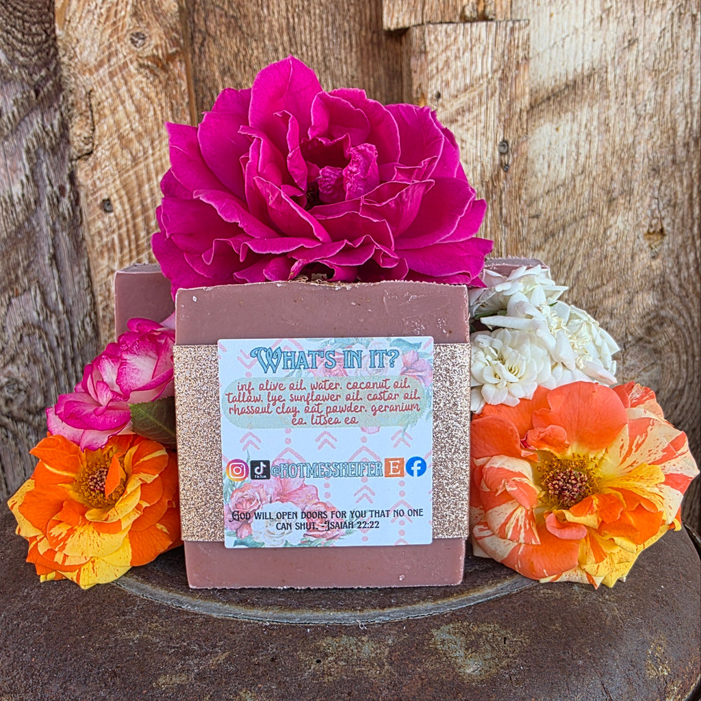A maroon-colored, handmade, beef tallow soap bar wrapped in a sparkly paper band and a decorative paper label, styled with bright roses on a rustic metal plate over a wooden surface. The label shows the ingredient list, social media handles, and a Bible verse.