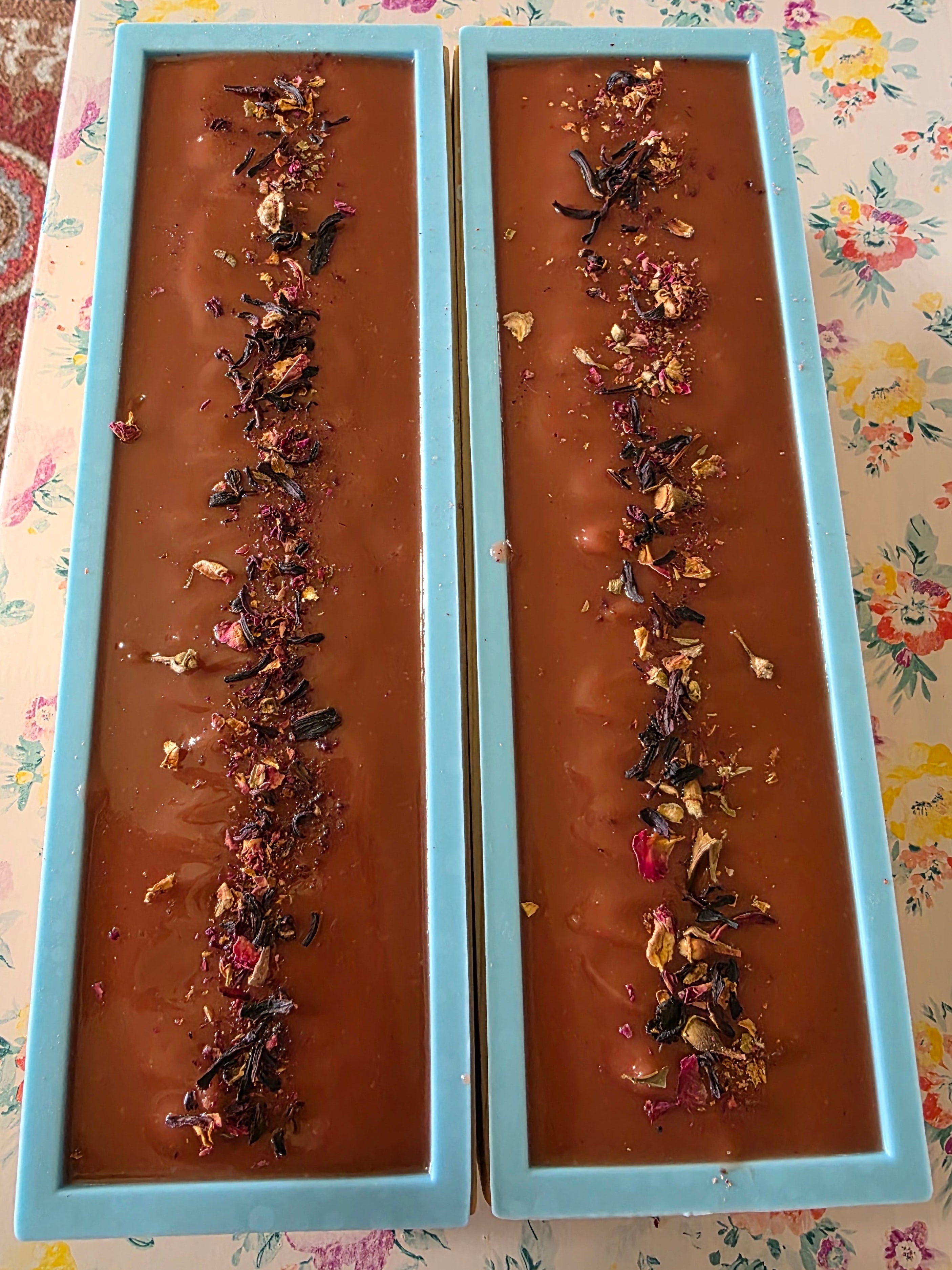 Two rectangular light blue molds filled with maroon-colored beef tallow soap, each topped with a decorative line of dried flower petals and botanicals, placed on a floral tablecloth.