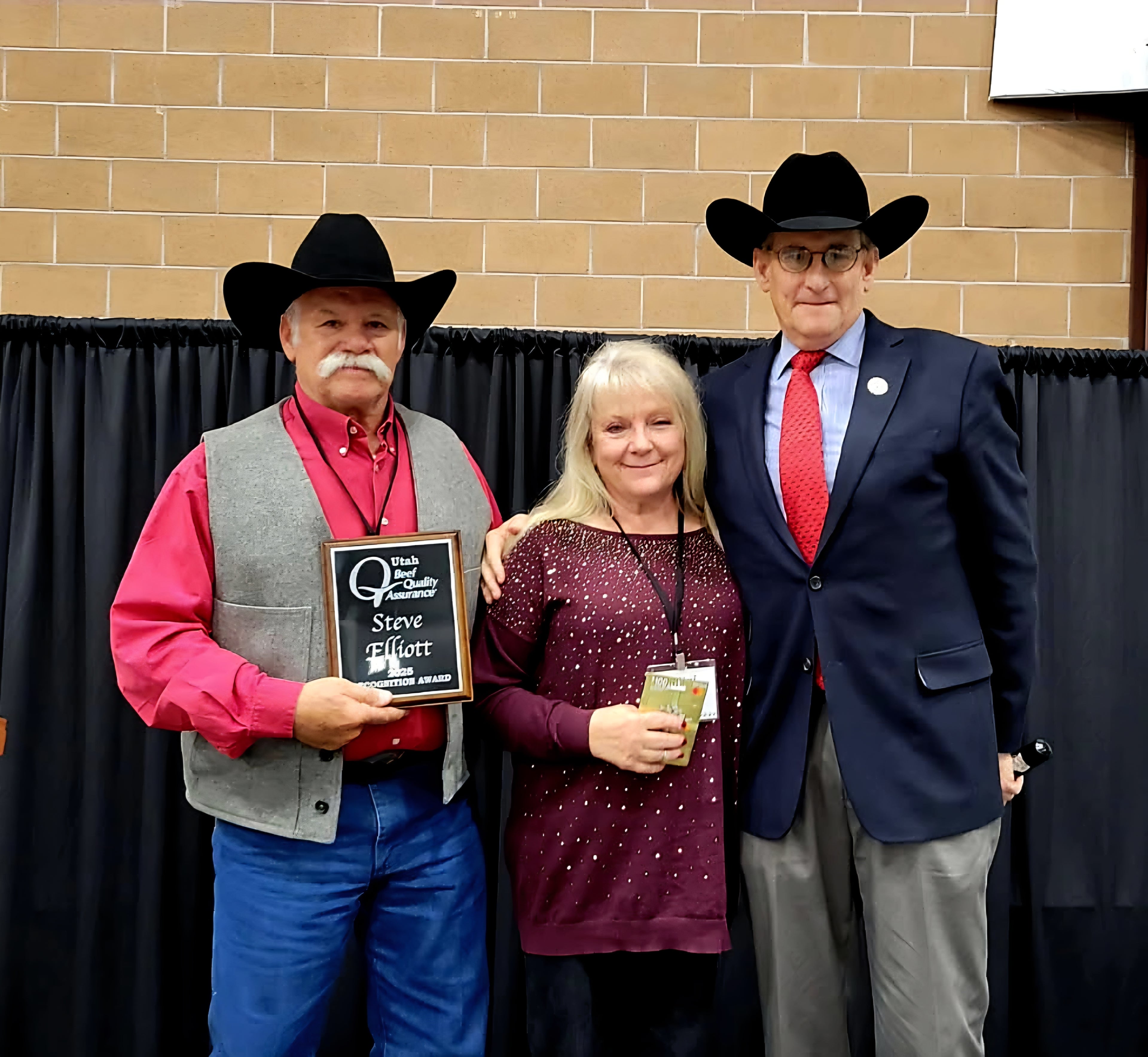 Beef cow rancher, his wife, and their friend posing together with a plaque against a brick wall.